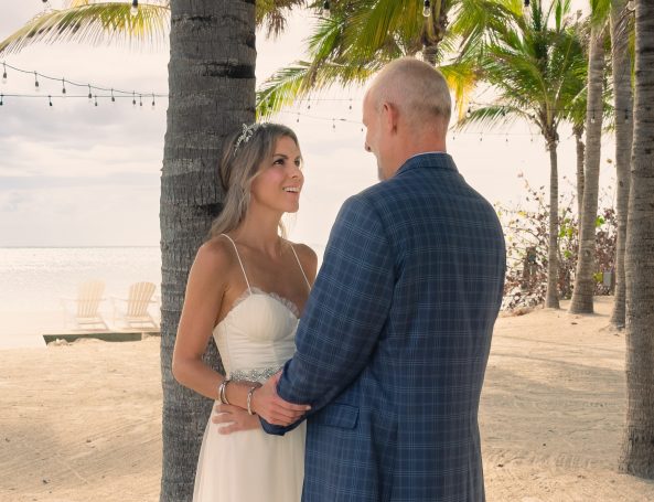 A bride and groom hold hands, smiling at each other under palm trees by the beach.