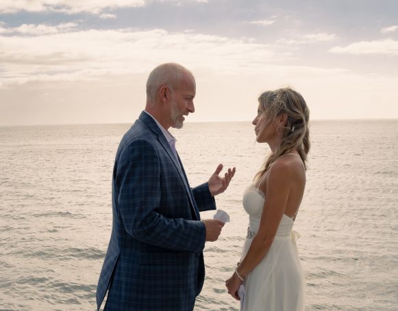 A couple in formal attire stands on a beach, smiling at each other with the ocean behind them.