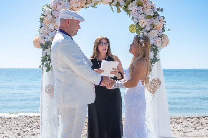 Couple exchanging vows under a floral arch on a beach. Officiant stands nearby.