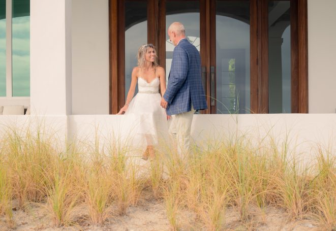 A bride and groom stand together, surrounded by tall grass near a beach house.