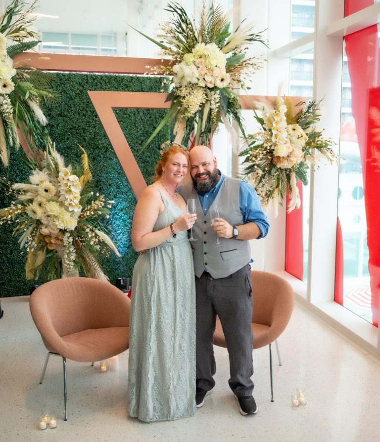 Bride & groom celebrating their wedding, posing in front of floral decorations and greenery backdrop before going on Virgin Cruise.