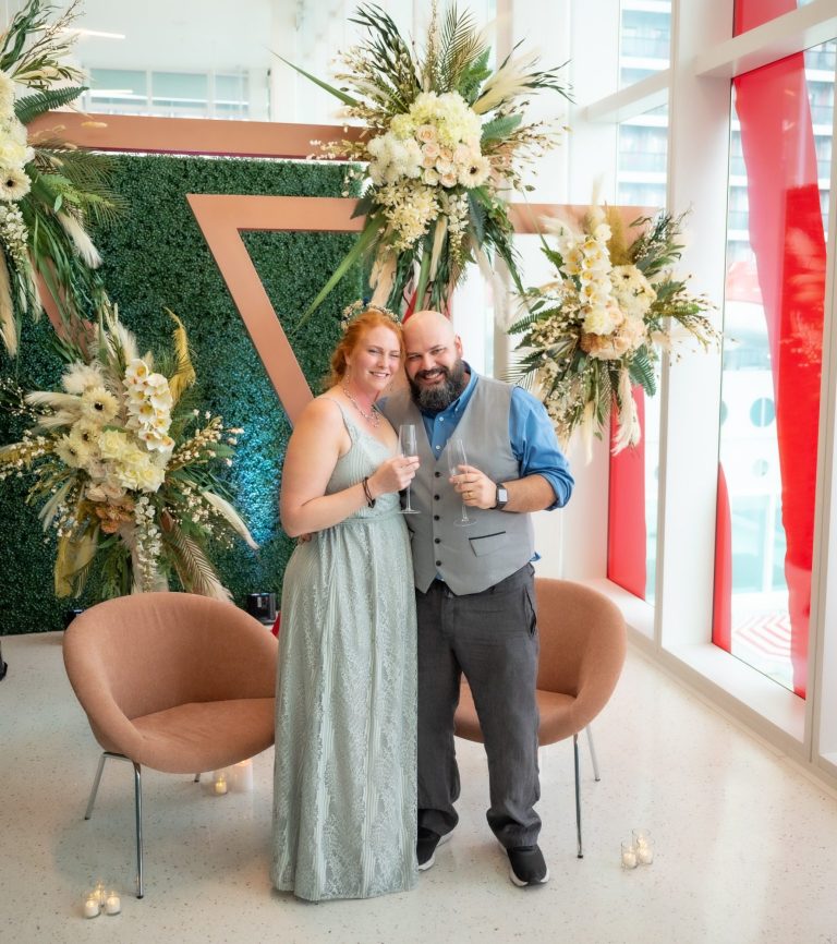 Bride & groom celebrating their wedding, posing in front of floral decorations and greenery backdrop before going on Virgin Cruise.