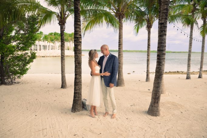 A couple embracing on a sandy beach near palm trees and calm waters.