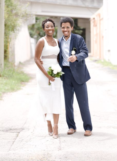 Smiling couple celebrating, dressed in formal attire, holding flowers in a lively outdoor setting.