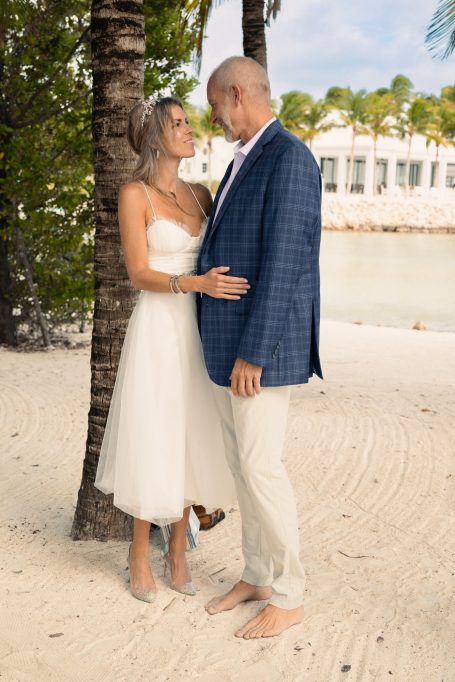 A couple embraces on a beach, dressed for a romantic occasion amidst palm trees.